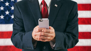 A politician is texting his staff during his political campaign, with the American flag as his backdrop.