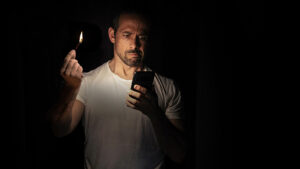 A man standing in the dark with a lit match in one hand and a mobile phone in the other receiving an emergency text alert. 