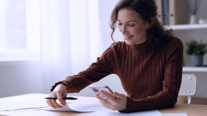 A woman sitting at a desk with her phone in her hand writing a brief sales text. 