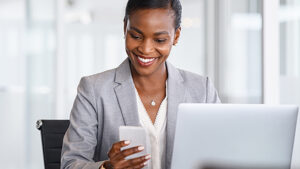 A woman sitting at a desk  with a computer in front of her looking at the mobile phone in her hand analyzing and adjusting her next sales text. 