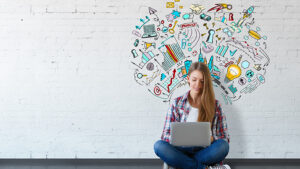 A woman is sitting with her laptop with a creative drawing on the wall behind her coming up with creative marketing ideas. 
